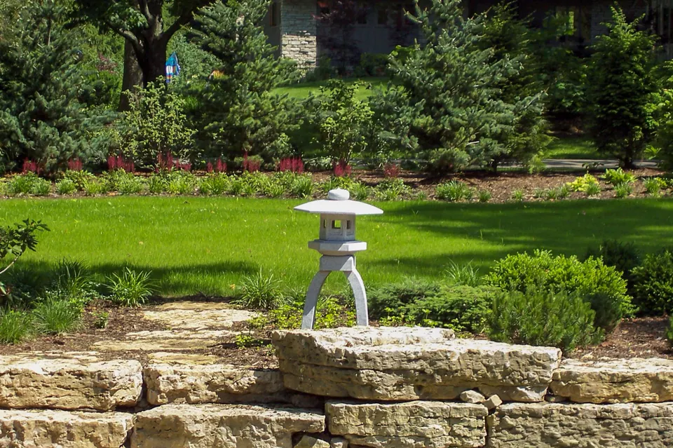 Stone lantern on stacked stone wall with lawn, clipped shrubs, red flowers, and evergreen backdrop.